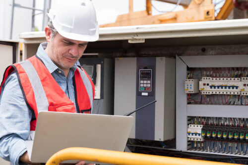 Electrician using laptop computer checking, repair, maintenance operation electric system at construction site with control panel